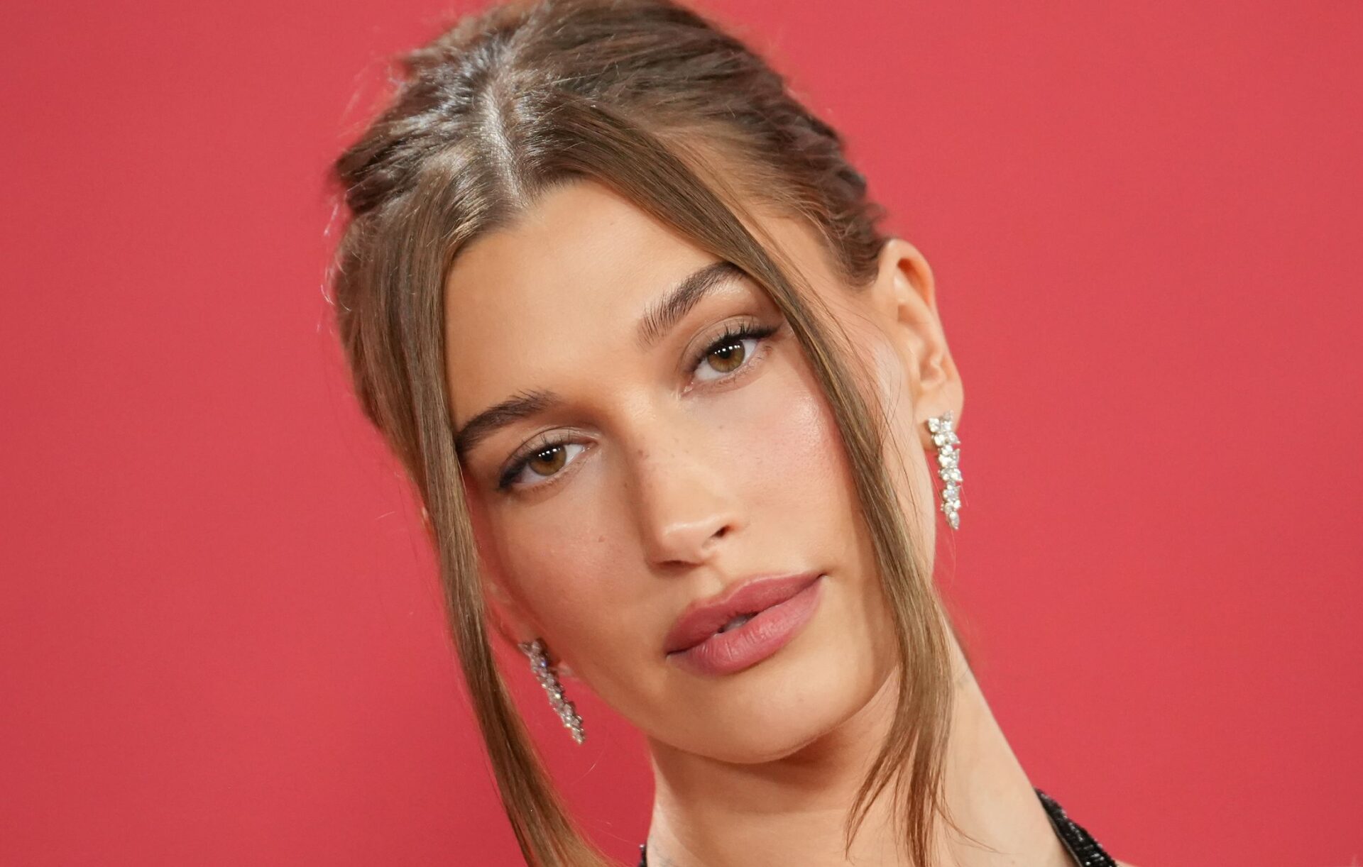 Hailey Bieber wears soft glam makeup and a slicked-back updo with face-framing pieces against a red backdrop